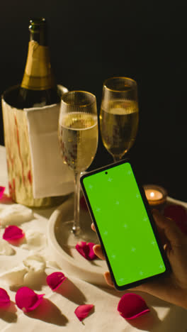 Vertical-Video-Of-Table-Set-For-Romantic-Marriage-Proposal-With-Champagne-And-Engagement-Ring-As-Person-Looks-At-Green-Screen-Phone